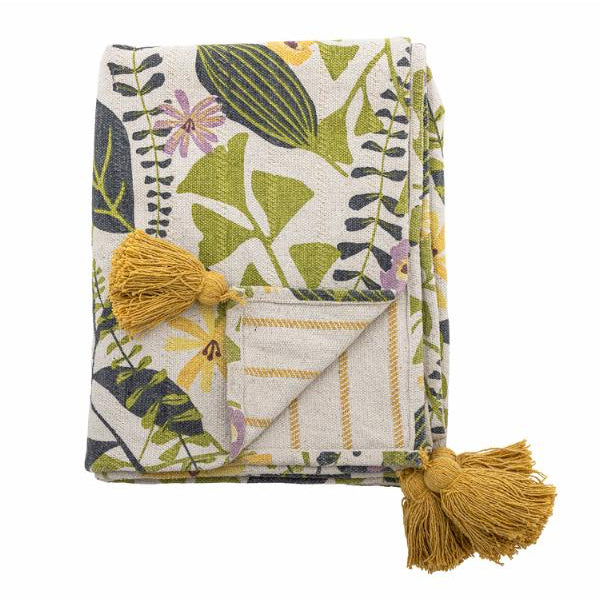 Een opgevouwen plaid met een botanisch bladmotief in groen, geel en paars op een crèmekleurige achtergrond, met twee mosterdgele kwastjes
