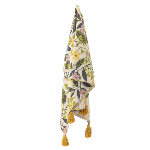 Een opgevouwen plaid met patroon en gele kwastjes, met een botanische print op een crèmekleurige achtergrond