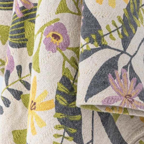 Een close-up van een opgevouwen geweven plaid met een botanische bloemenprint op een crèmekleurige achtergrond, met groene bladeren, paarse bloemen en gele accenten.