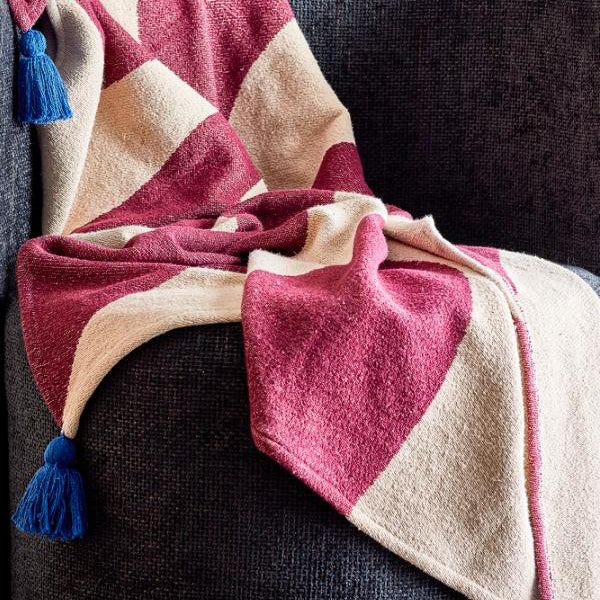 Une couverture douce à grandes rayures diagonales rose poudré et crème, ornée de pompons bleus, drapée sur un fauteuil gris foncé texturé.