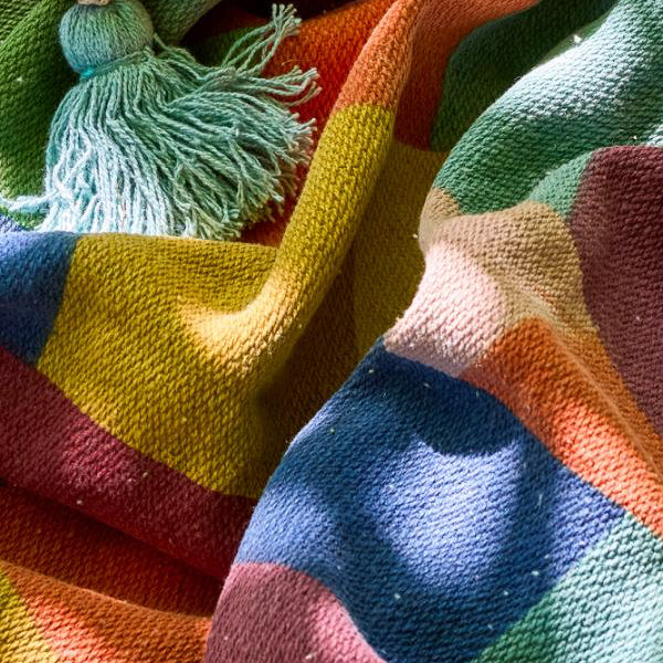A close-up of a colourful woven plaid blanket with a tassel, showing bold blocks of orange, green, yellow, blue, pink, and red in bright sunlight.