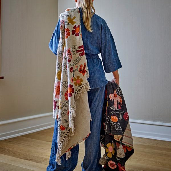 A woman stands with her back to the camera, holding up two patterned plaids. One throw is cream with orange, red, and green floral motifs, and the other is black with a multicolor floral print.