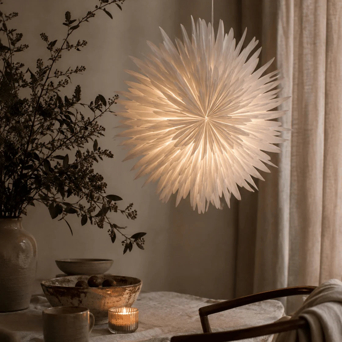 A glowing white paper spiky pendant light hangs in a softly lit room beside a table with bowls, a candle, and a vase of branches.