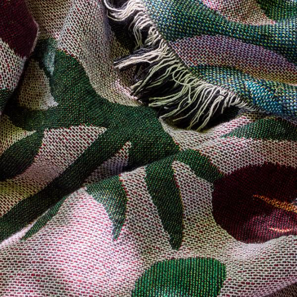 A close-up of a woven textile with a botanical pattern in green, burgundy, and lavender on a pale pink base, with fringed edges visible near the top.