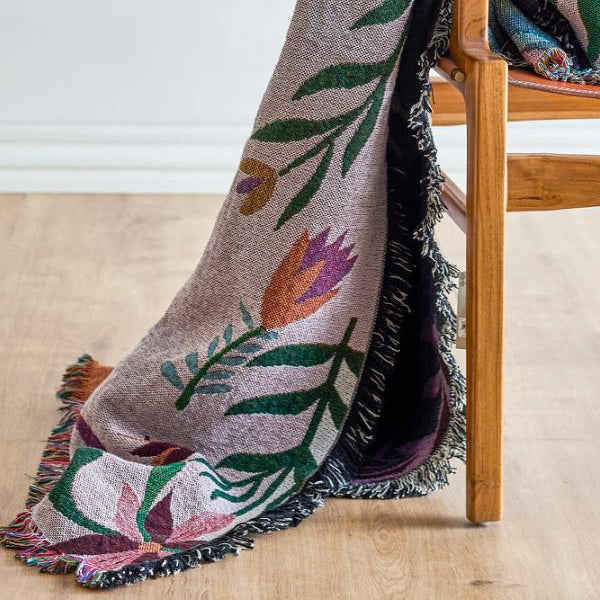 A woven throw blanket with a floral tulip pattern draped over a wooden chair in a bright room. The blanket has a light taupe-beige base with green leaves, pink and purple tulips, and black fringe.