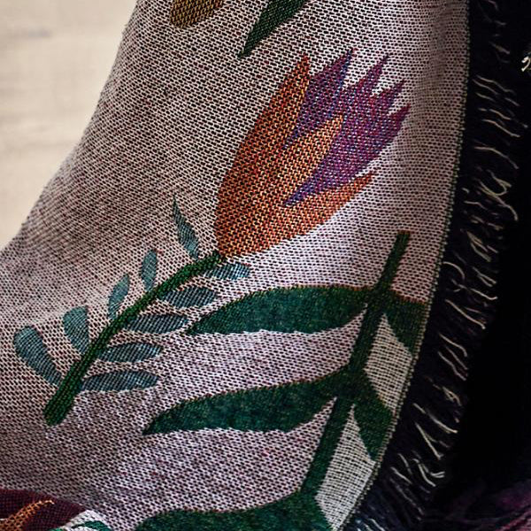 A close-up of a woven textile with a floral pattern in muted green, orange, purple, and pink on a taupe background, with fringe visible along one edge.