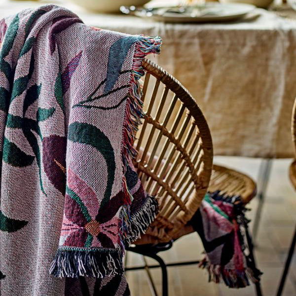 A woven throw blanket with a pink, purple, and green floral pattern and fringed edges draped over a wicker chair in a softly lit dining room setting.
