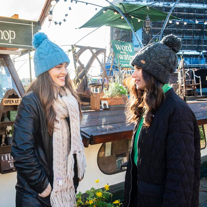 Two women standing and smiling in front of a bookshop kiosk on a boat, wearing winter knit beanies.