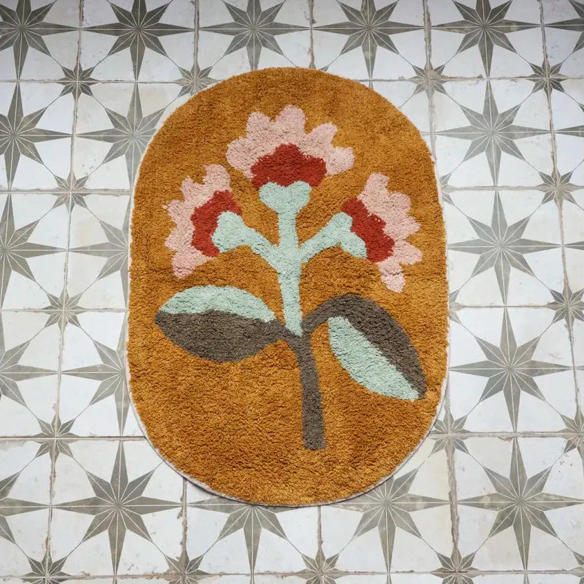 An oval bath mat with a stylized floral design, featuring a mustard brown background and pink, red, light green, and gray-green leaves by Madam Stoltz