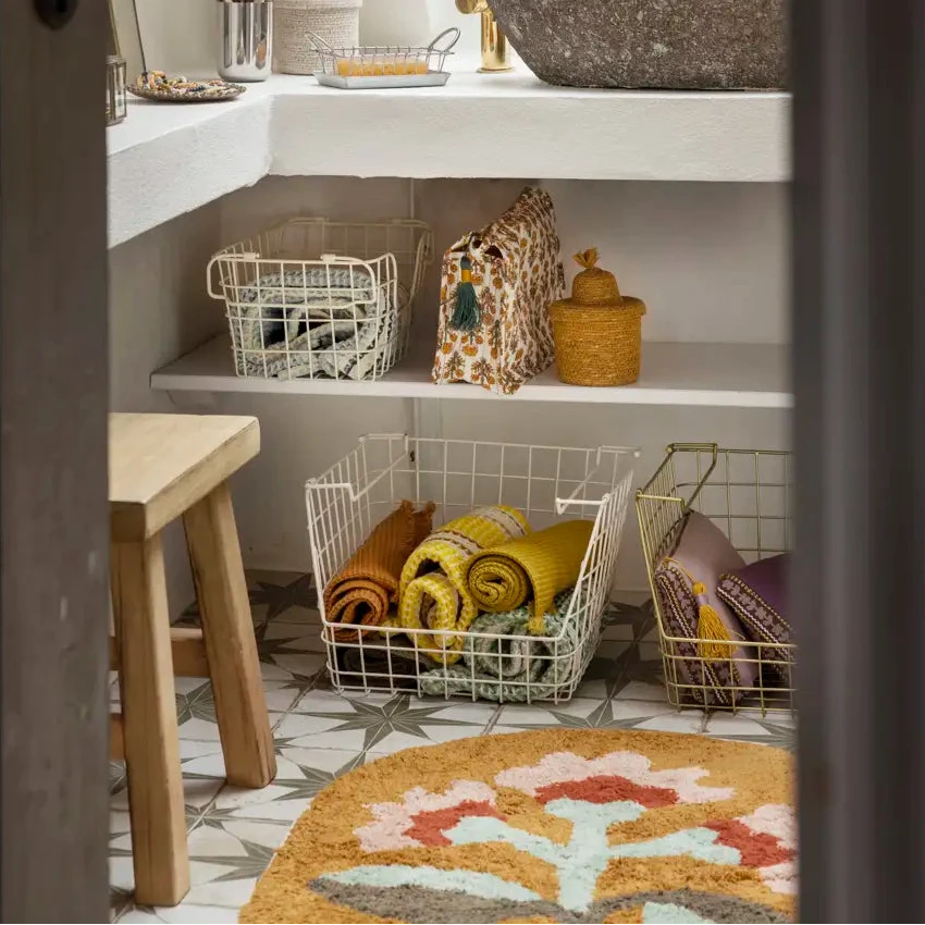 A styled bathroom interior with a white L-shaped vanity, a dark stone vessel sink, a gold faucet, open shelving with wire baskets, and a patterned bath mat