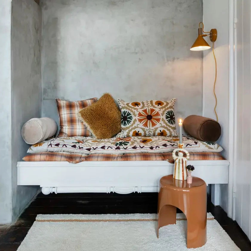 A rustic bohemian-style daybed with patterned plaid and textured pillows, set in a minimalist room with a concrete wall, white ceiling, and a small wooden stool.
