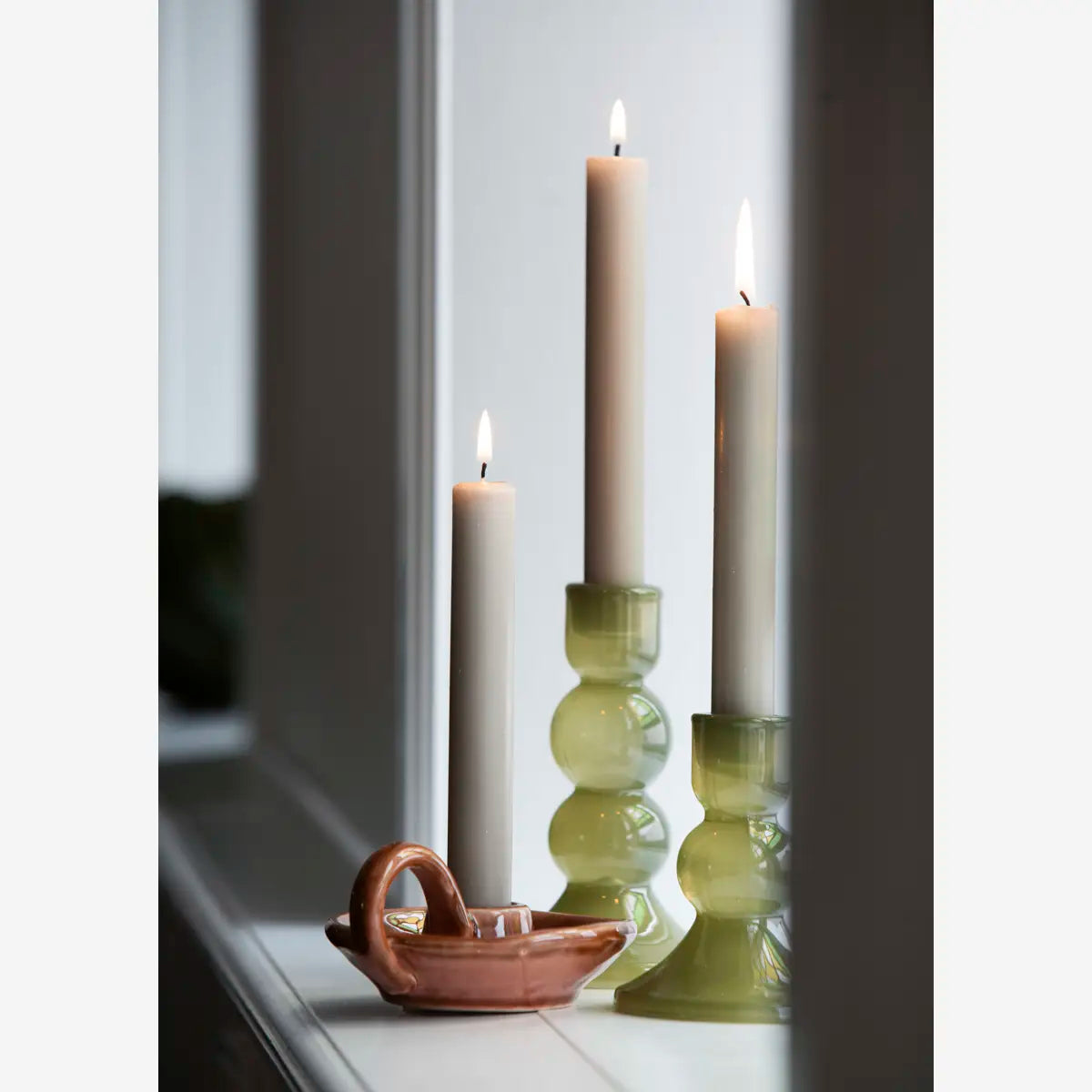 Three lit taper candles in green glass candlesticks and a small terracotta boat-shaped holder on a windowsill.