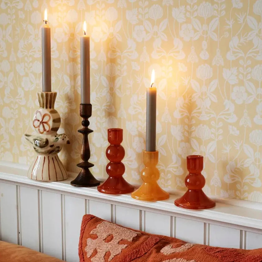 A styled interior scene with four lit taper candles in decorative holders arranged on a white mantel against patterned wallpaper, with part of a rust-colored sofa and textured pillow visible in the foreground.