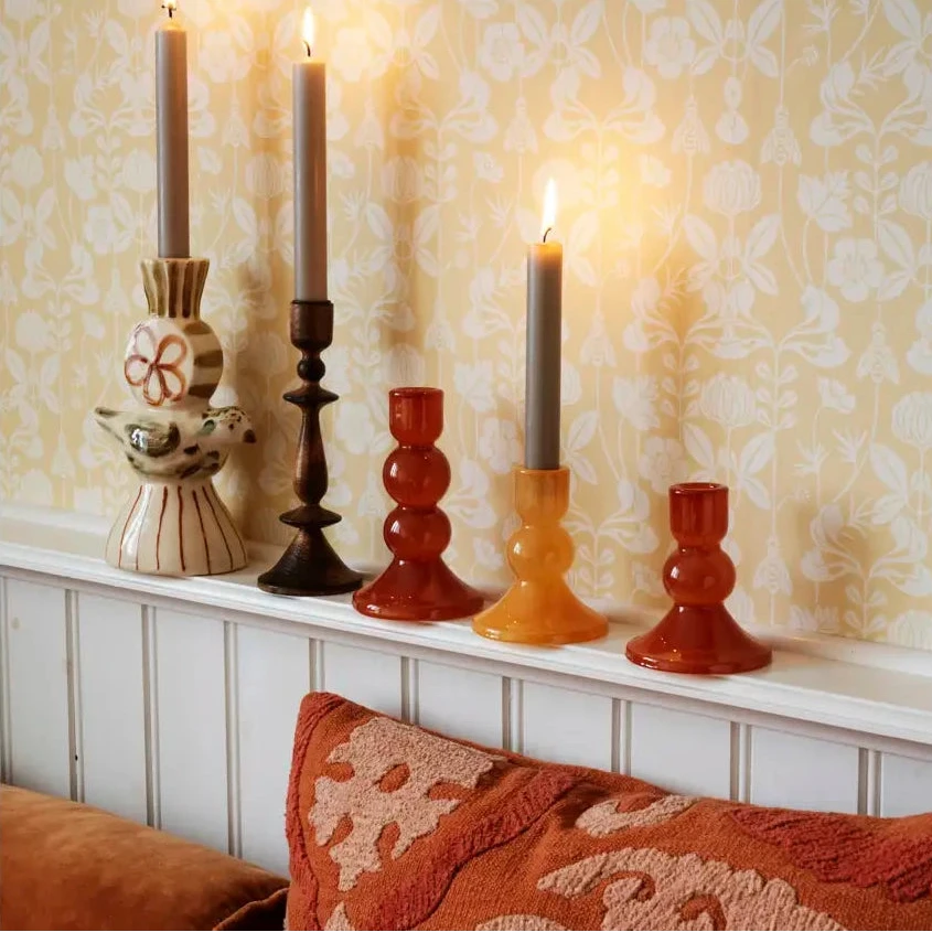 A styled interior scene with four lit dinner candles in glass holders with part of a rust-coloured sofa and textured pillow visible in the foreground.