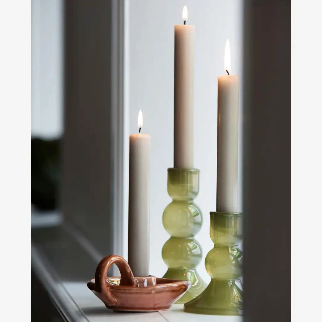 Three lit dinner candles in green glass candlesticks and a small terracotta boat-shaped holder on a windowsill.