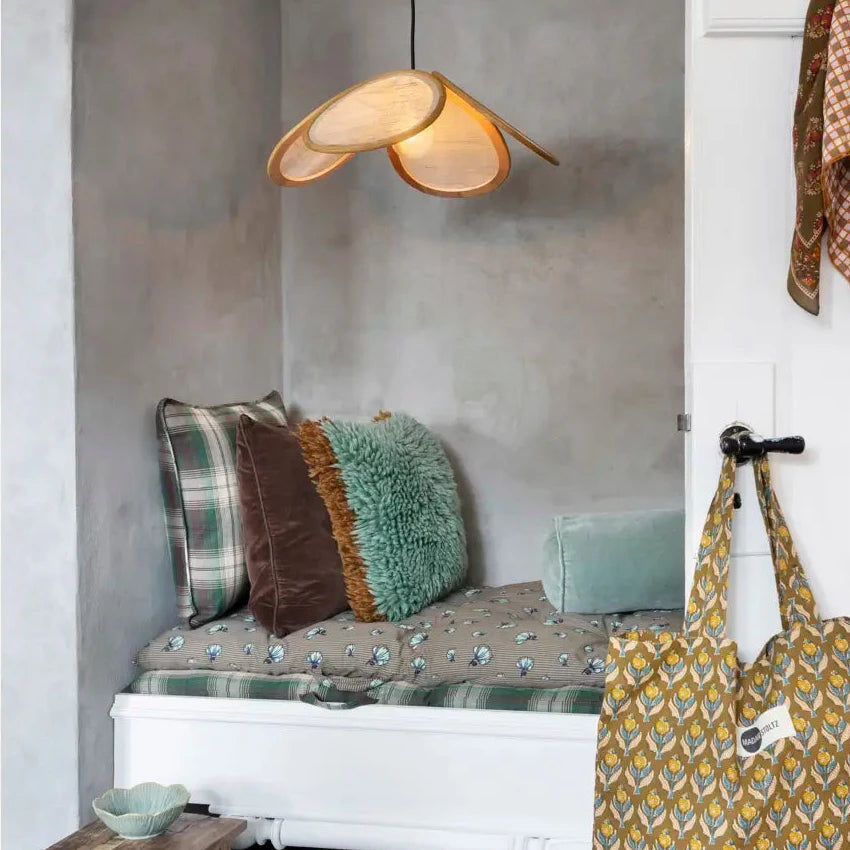 A styled interior corner with a white bench against a gray plaster wall, patterned pillows, a hanging woven bag on a door hook, a small wooden stool, and a pendant light.