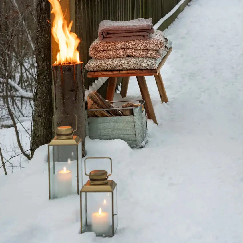 Outdoor winter scene with a snow-covered walkway beside a wooden fence and tree, featuring a lit fire in a metal barrel, folded blankets on a wooden bench, lanterns with candles.