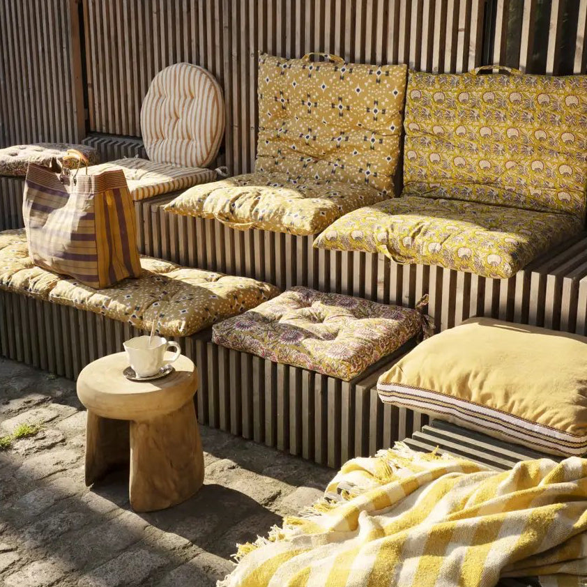 Outdoor seating area with yellow cushioned sofa and ottoman, wooden stool, and striped wall.