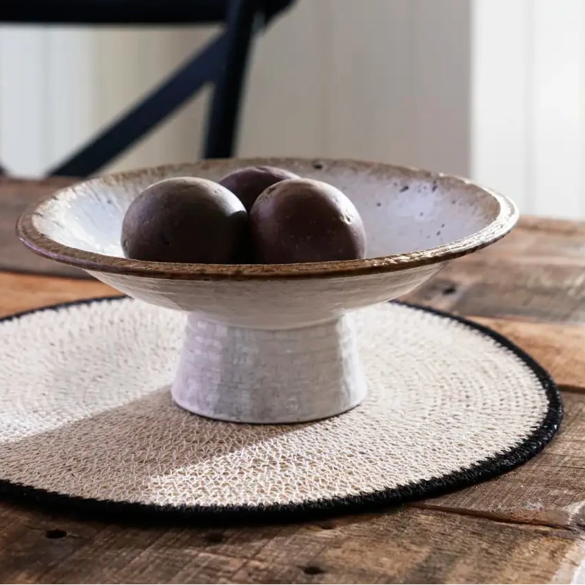 A ceramic pedestal bowl set on a woven placemat on a wooden table.