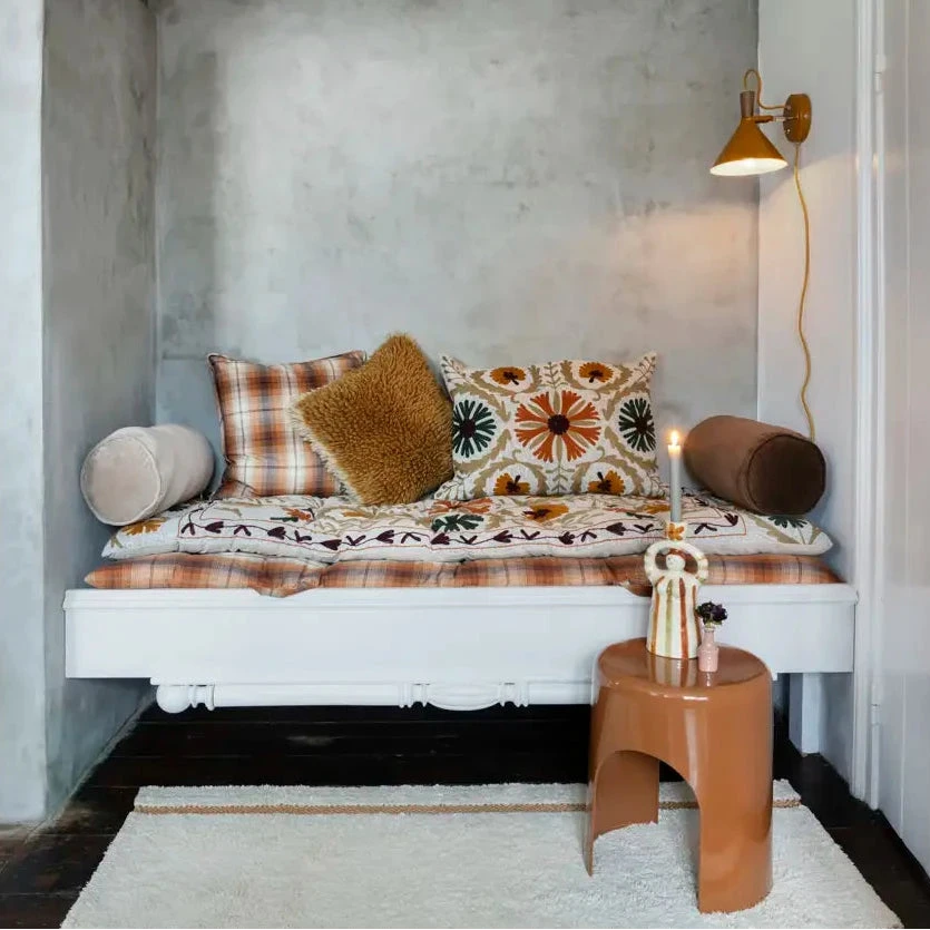 A rustic bohemian-style daybed with patterned plaid and textured pillows, set in a minimalist room with a concrete wall, white ceiling, and a small wooden stool.