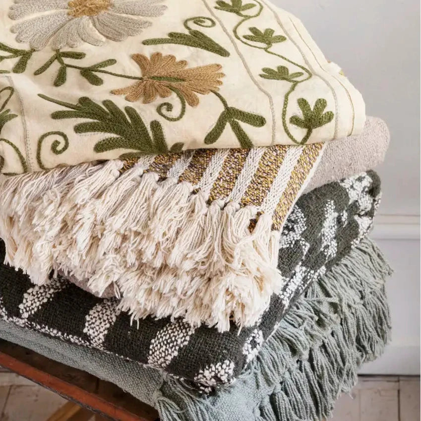 Stack of decorative blankets with floral patterns and fringes on a wooden chair.