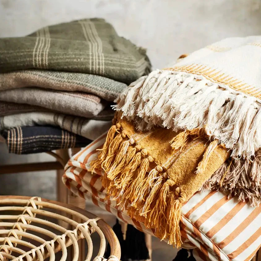Madam Stoltz styled arrangement of folded and draped textiles, including a mustard yellow fringed throw, a cream and tan striped pillow, a green plaid blanket, and woven rattan furniture.