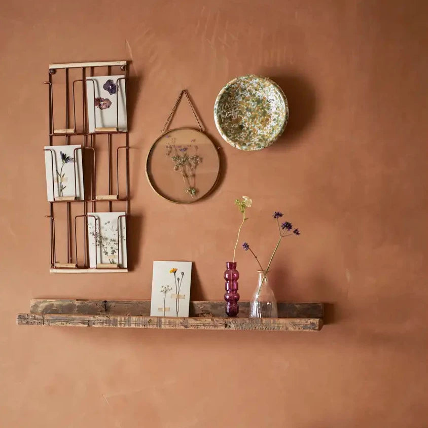 Decorative wall shelf with a vase, small plant, and framed pictures against a terracotta wall.