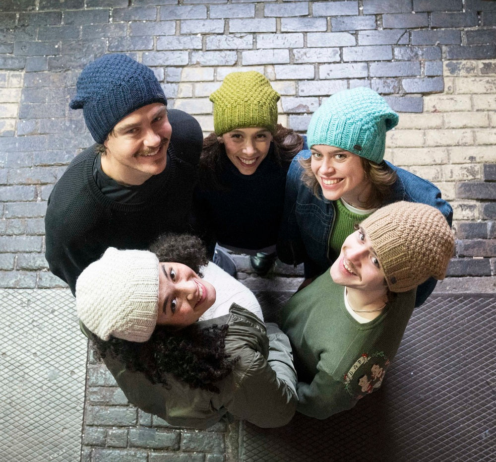 Five people gathered closely together,  each wearing a different knit beanie in navy, yellow, light blue, cream, and tan.