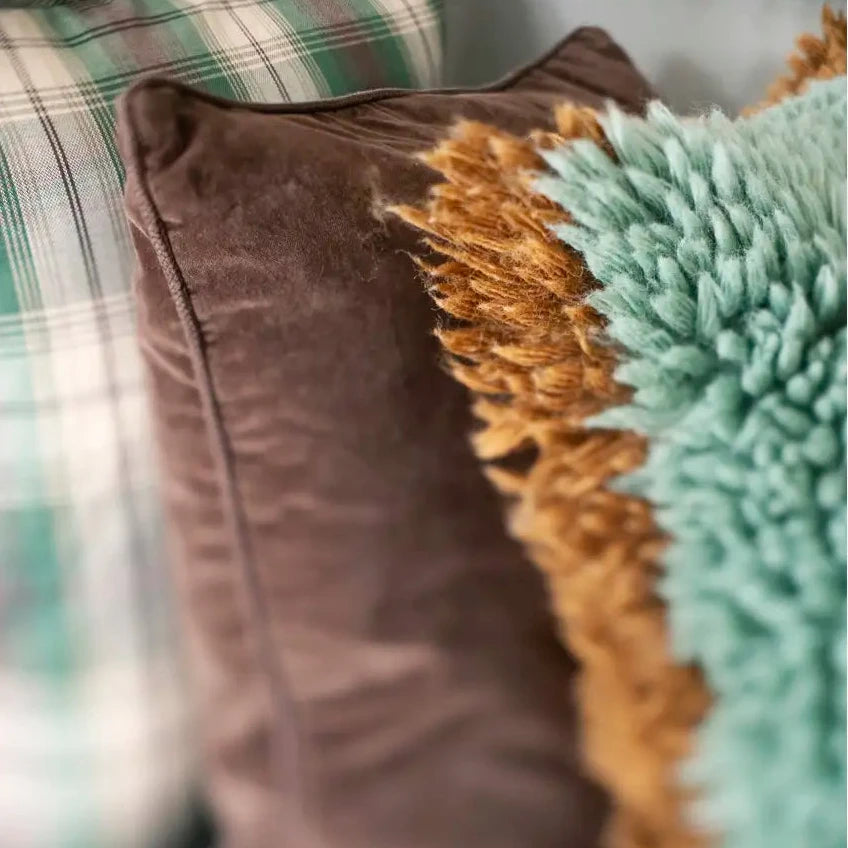 A close-up of decorative throw pillows, featuring a prominent brown velvet pillow in the center, a mint green shaggy pillow with a tan fringe