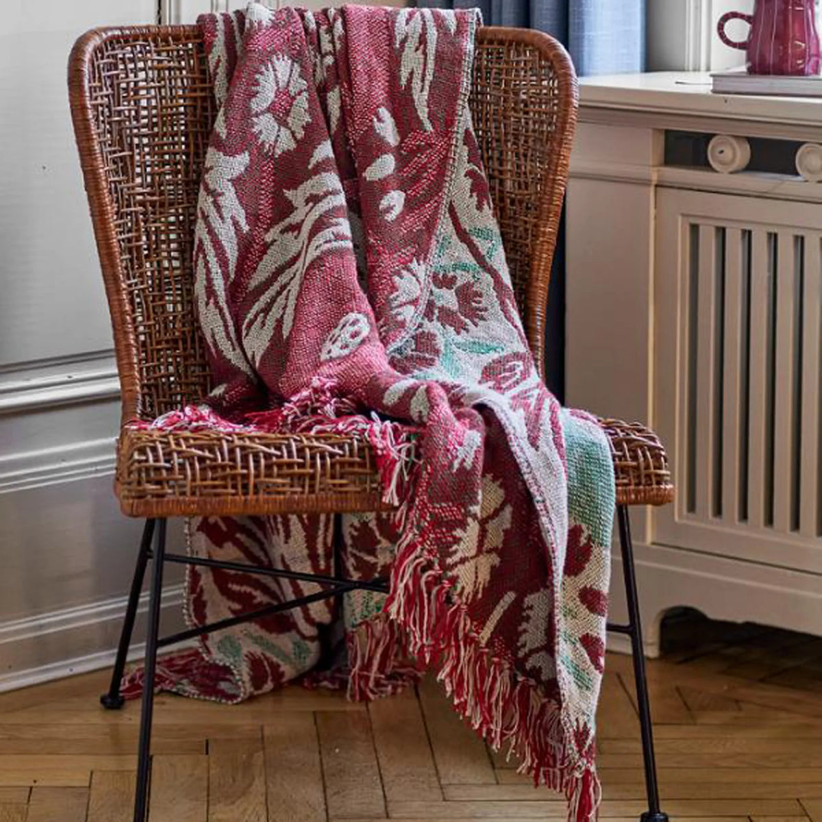 A woven wicker chair in a home setting with a red, cream, and mint green patterned throw blanket draped over it.