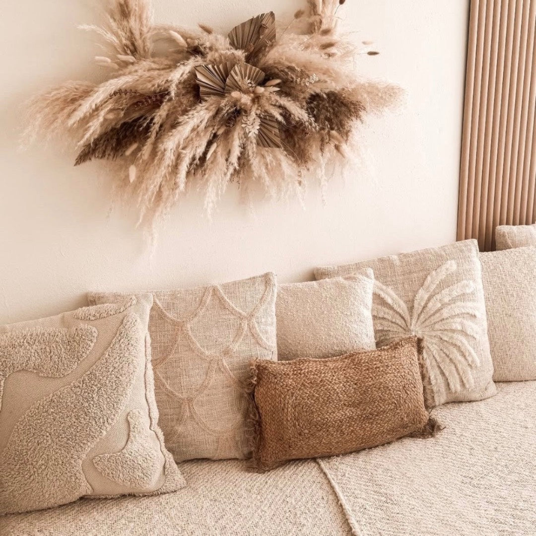 A styled bohemian home decor scene featuring a beige sofa adorned with textured throw pillows, a brown jute accent pillow, and a dried pampas grass wall hanging on a cream wall.