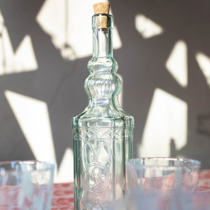 A clear glass bottle with an embossed pattern and a cork stopper, set on a table with two glasses in the foreground