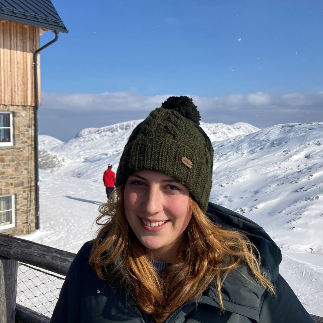 A smiling woman outdoors in a snowy mountain setting, wearing an olive green knit pom-pom beanie and a dark winter jacket.