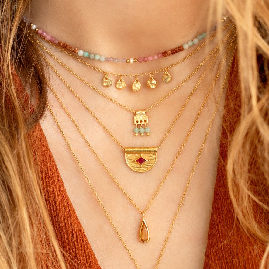 A close-up of a person wearing multiple layered gold necklaces, including a beaded choker, a semicircle pendant with a red stone, a teardrop pendant, and a larger ornate pendant with a red stone.