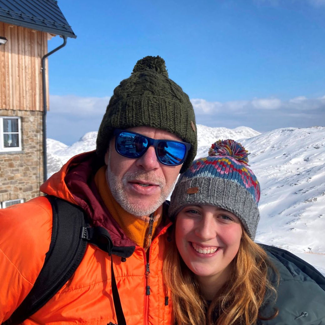 A man and a woman taking a selfie in a snowy mountain setting. The man wears an orange jacket, a dark green knit beanie, and blue mirrored sunglasses, while the woman wears a gray knit beanie with a multicolor pom-pom.
