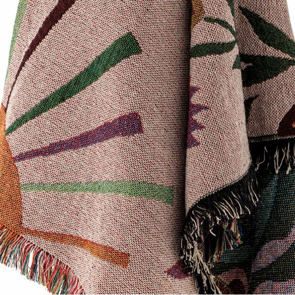 A close-up of a woven throw blanket with fringe, featuring a beige base with abstract sunburst and leaf-like patterns in green, purple, orange, and dark teal.